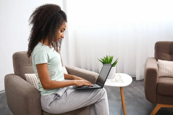 Beautiful African-American woman with laptop sitting in armchair at home — Stock Photo, Image