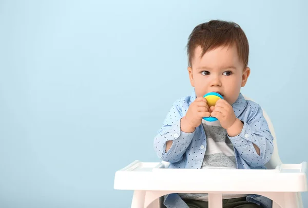 Cute little boy with nibbler sitting in high chair against color background — Stock Photo, Image