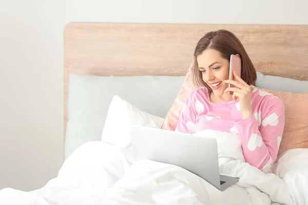 Morning of beautiful young woman talking by phone while working on laptop in bed — Stock Photo, Image