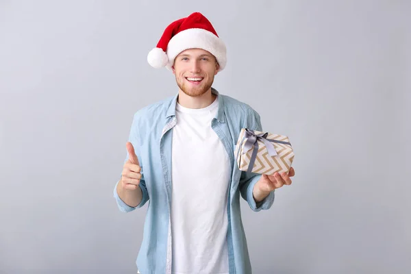 Young man with Christmas gift box showing thumb-up on light background — Stock Photo, Image