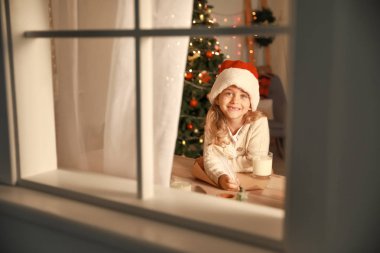 Noel arifesinde Noel Baba'ya evde mektup yazan sevimli küçük kız. Pencereden görüntüle