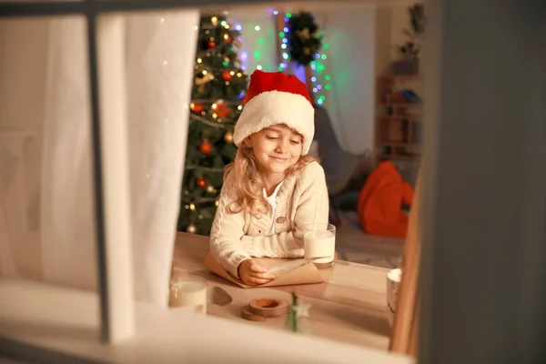 Cute Little Girl piśmie list do Świętego Mikołaja w Wigilię Bożego Narodzenia w domu. Widok przez okno — Zdjęcie stockowe