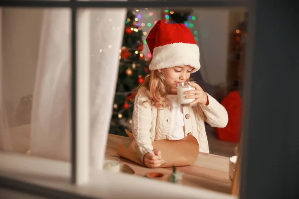 Cute little girl writing letter to Santa Claus and drinking milk on Christmas eve at home. View through window — Stock Photo, Image