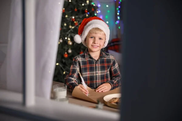 Mignon petit garçon qui écrit une lettre au Père Noël la veille de Noël à la maison. Vue par fenêtre — Photo