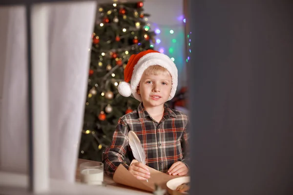 Mignon petit garçon qui écrit une lettre au Père Noël la veille de Noël à la maison. Vue par fenêtre — Photo