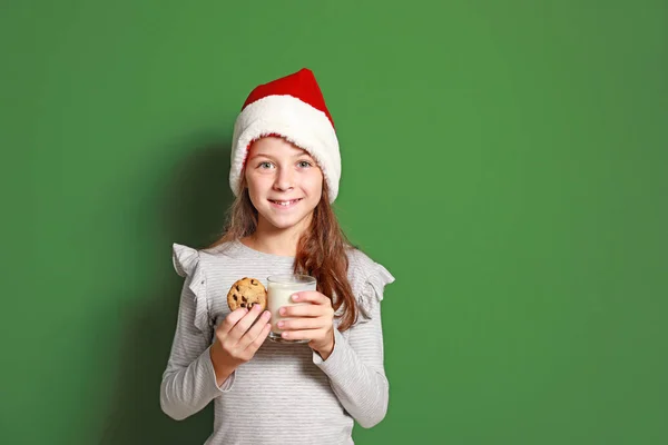 Menina bonito em chapéu de Papai Noel, com leite e biscoito no fundo de cor — Fotografia de Stock