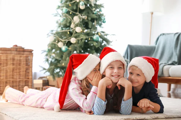 Gossiping crianças pequenas em chapéus de Papai Noel deitado no chão em casa — Fotografia de Stock