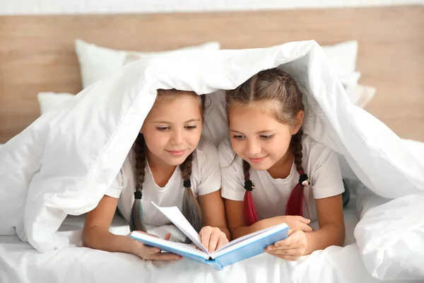 Portrait of twin girls reading book under blanket on bed — Stock Photo, Image