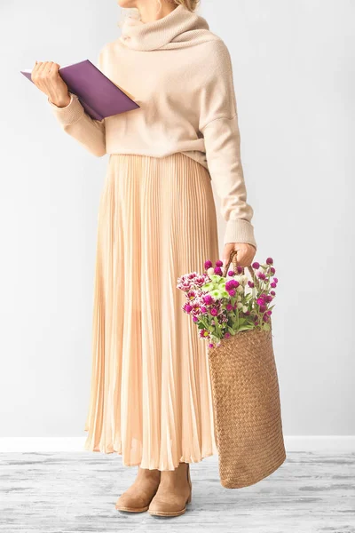 Женщина с книгой и цветами в эко-сумке возле светлой стены — стоковое фото