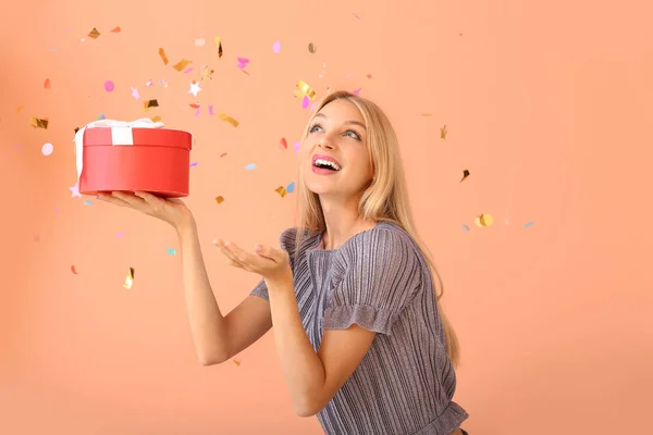 Hermosa mujer joven con regalo y confeti cayendo sobre fondo de color —  Fotos de Stock