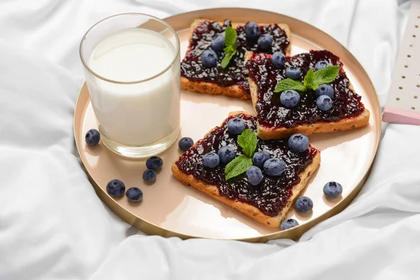 Plate with toasted bread, berry jam and glass of milk on bed — Stock Photo, Image
