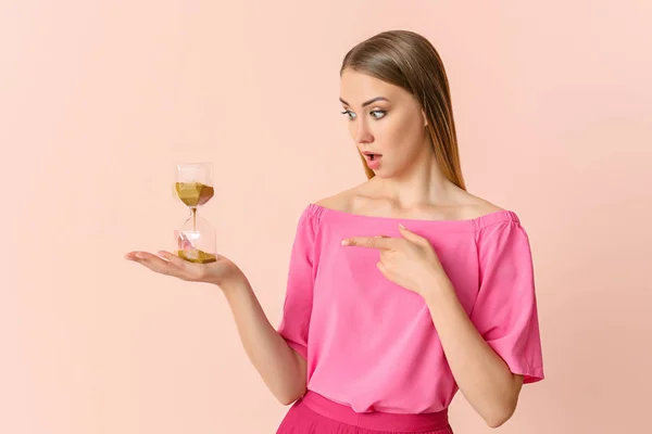 Shocked young woman with hourglass on color background — Stock Photo, Image