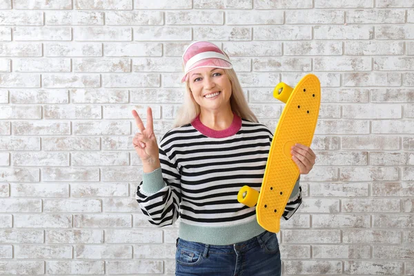 Stylish mature woman with skateboard showing victory gesture on brick background — Stock Photo, Image
