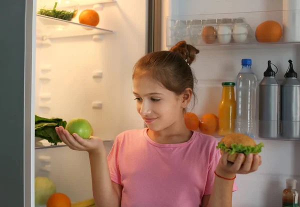 Девушка со здоровой и нездоровой пищей возле холодильника ночью — стоковое фото