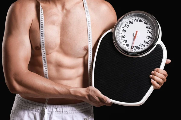 Handsome muscular man with measuring tape and scales on dark background. Weight loss concept — Stock Photo, Image