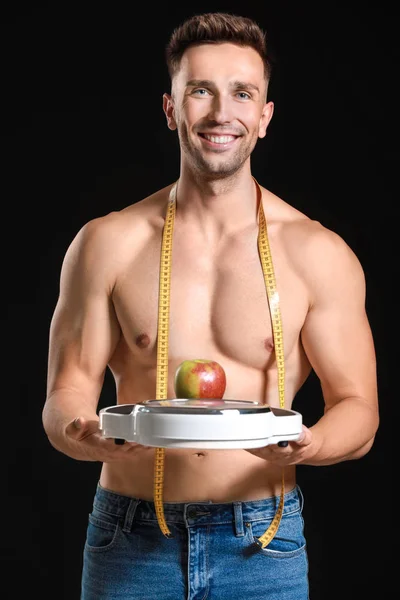 Όμορφος μυώδης άνθρωπος με μέτρηση ταινία, κλίμακες και μήλο σε σκούρο φόντο. Έννοια απώλειας βάρους — Φωτογραφία Αρχείου