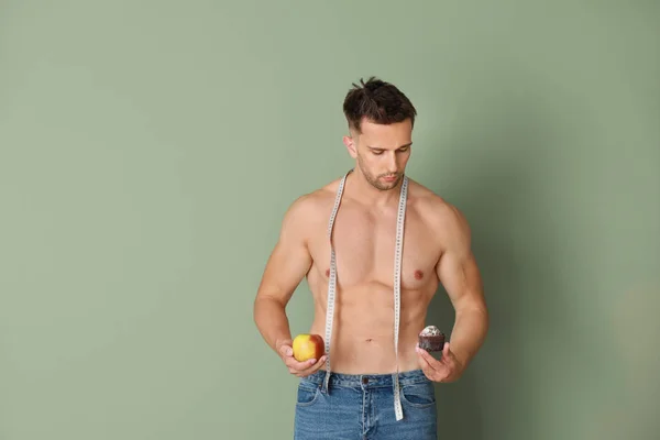 Handsome muscular man with measuring tape, dessert and apple on color background. Weight loss concept — Stock Photo, Image