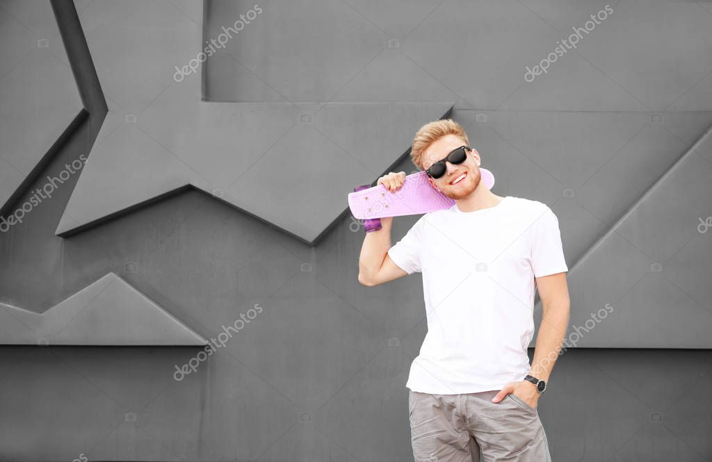 Man in stylish t-shirt and with skateboard near grey wall