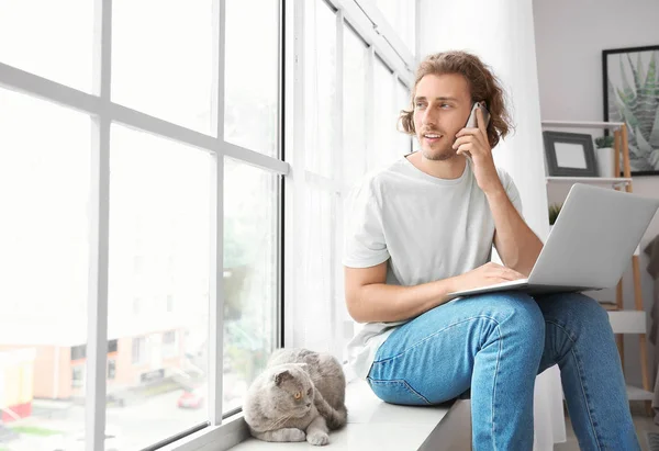 Man with laptop talking by phone while sitting on window sill with cute cat — Stock Photo, Image