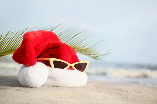 Kapelusz Świętego Mikołaja z okulary przeciwsłoneczne i tropikalny liść na wybrzeżu morza. Świąteczne wakacje — Zdjęcie stockowe