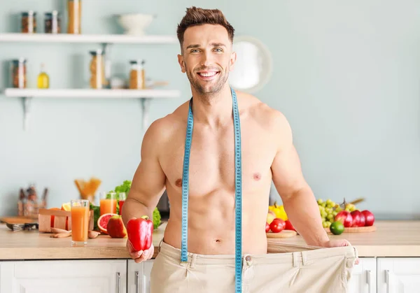 Handsome muscular man in loose pants, with measuring tape and pepper in kitchen. Weight loss concept