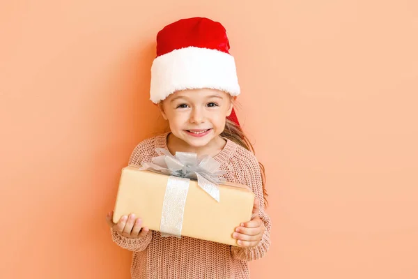 Kleines Mädchen mit Weihnachtsmütze und Weihnachtsgeschenk auf farbigem Hintergrund — Stockfoto