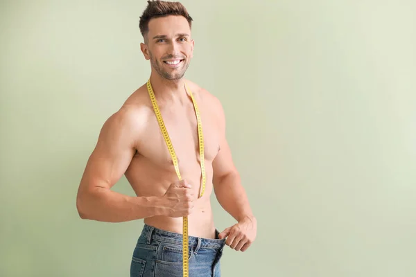 Schöner muskulöser Mann in lockeren Jeans und mit Maßband auf farbigem Hintergrund. Gewichtsverlust-Konzept — Stockfoto