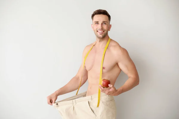 Handsome muscular man in loose pants, with measuring tape and apple on light background. Weight loss concept