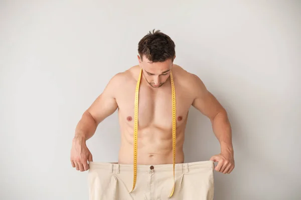 Handsome muscular man in loose pants and with measuring tape on light background. Weight loss concept — Stock Photo, Image