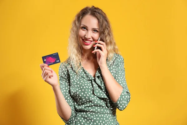 Young woman with credit card talking by mobile phone on color background — Stock Photo, Image