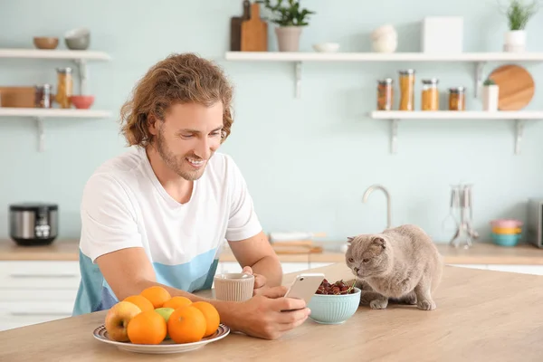 Man with mobile phone drinking coffee while sitting in kitchen with cute cat — Stock Photo, Image