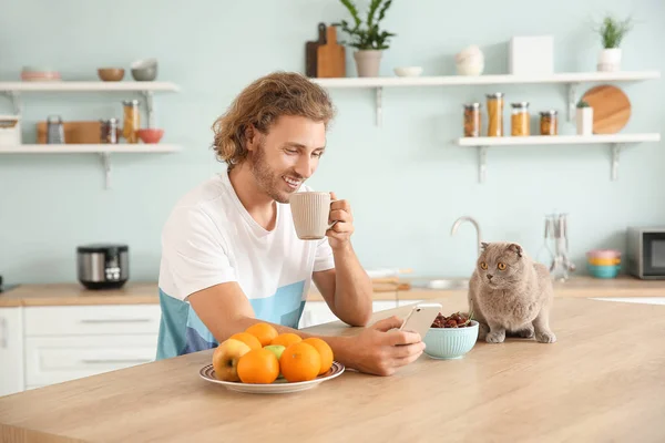 Man with mobile phone drinking coffee while sitting in kitchen with cute cat — Stock Photo, Image