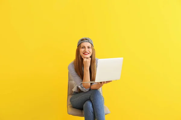 Happy young woman with laptop sitting on chair against color background — Stock Photo, Image