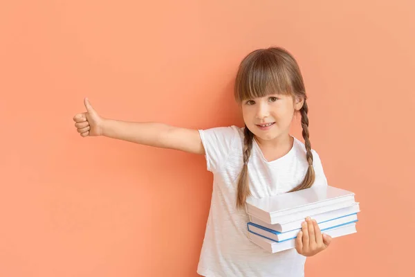 Cute little girl with books showing thumb-up on color background — Stock Photo, Image