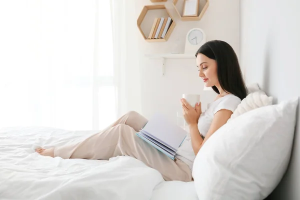 Beautiful young woman drinking coffee while reading book in bedroom — Stock Photo, Image