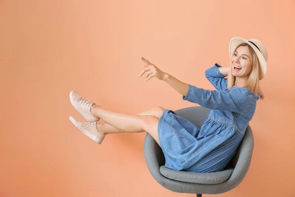 Stylish woman sitting in armchair and pointing at something on color background — Stock Photo, Image