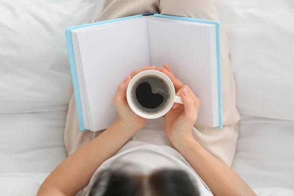 Beautiful young woman drinking coffee while reading book in bedroom, top view — Stock Photo, Image