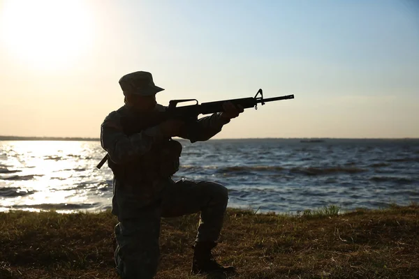 Silhouette of soldier in camouflage taking aim near river — Stock Photo, Image