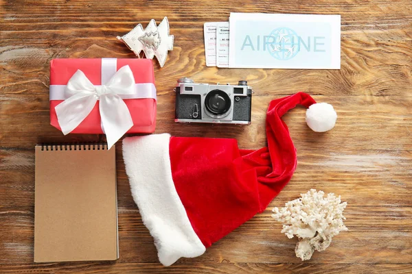 Composition with gift box, Santa Claus hat, photo camera and tickets on wooden background. Christmas vacation