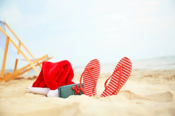 Santa Claus hoed met geschenk doos en Teenslippers op zee kust. Kerst vakantie — Stockfoto