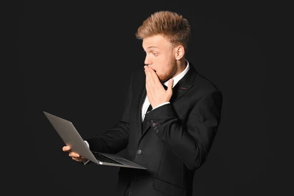 Shocked young businessman with laptop on dark background — Stock Photo, Image