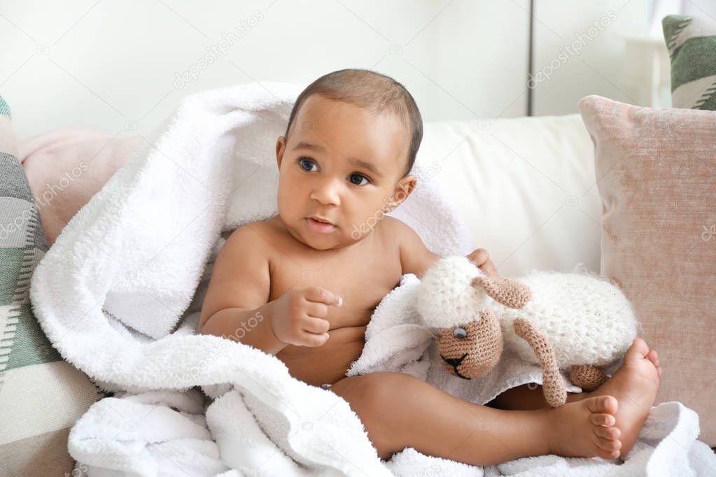 Cute African-American baby with soft towel and toy on bed at home