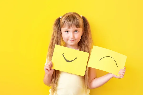 Little girl with drawn smiling and upset mouths on sheets of paper against color background — Stock Photo, Image