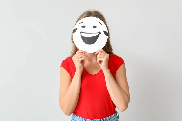 Young woman hiding face behind emoticon against light background — Stock Photo, Image