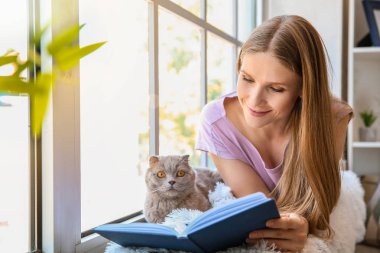 Evde pencere yakınında sevimli kedi okuma kitabı ile güzel kadın