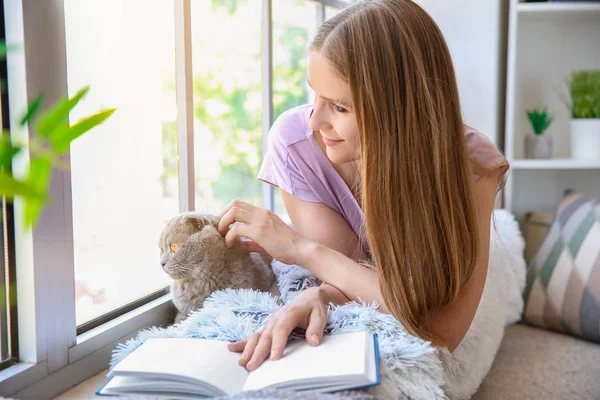 Beautiful woman with cute cat reading book near window at home — Stock Photo, Image