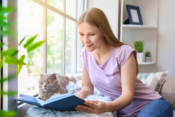 Женщина с симпатичной кошкой, читающей книгу у окна дома — стоковое фото