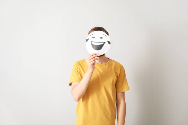 Man hiding face behind sheet of paper with drawn emoticon on grey background
