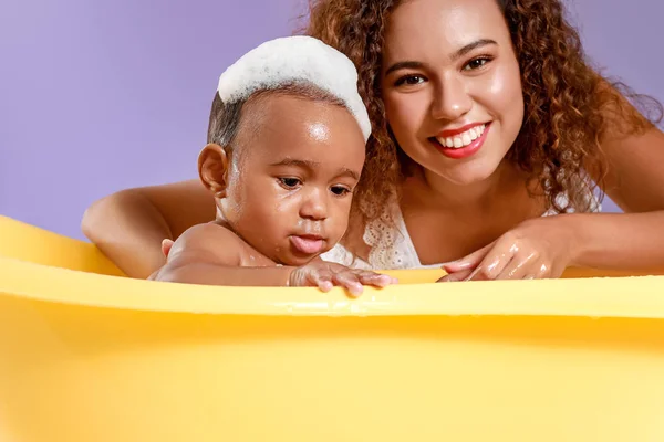 Молодая афро-американская мать моет своего ребенка в ванне на цветном фоне — стоковое фото