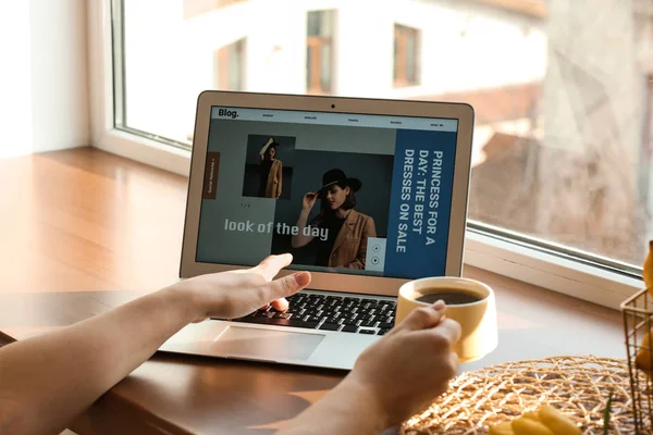Female fashion blogger working on laptop and drinking coffee near window — Stock Photo, Image